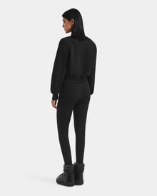 A woman with straight dark hair, wearing the Rudsak LIDDIE - L525505 Black cropped crewneck sweater, black leggings, and black platform boots, stands facing away against a plain white background.