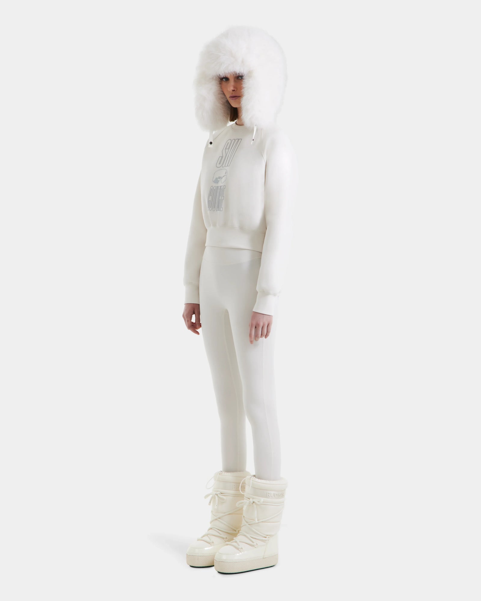 A person models the Rudsak LIDDIE - L525505 Vanilla: an all-white ski-inspired outfit with a furry hooded sweatshirt, fitted pants, and tall padded snow boots, effortlessly chic against a clean white backdrop.