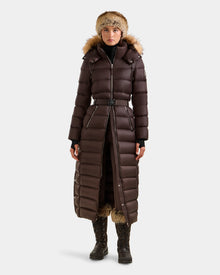 Wearing the Rudsak LILITH L - L125528 Espresso maxi down puffer with RDS-certified down, removable belt, fur-lined hood, plus a black turtleneck, gloves, winter boots, and a fur hat, a person stands against a plain light background.