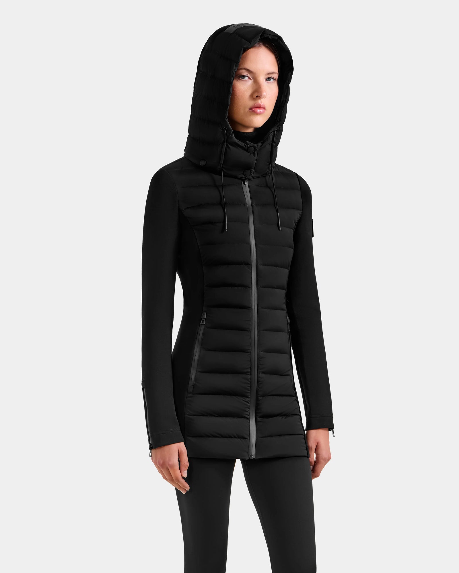 A woman wearing the Rudsak MARRY M - L125519 Black mid-length hybrid coat with a front zipper stands against a plain light gray background, hands relaxed at her sides and a neutral expression.