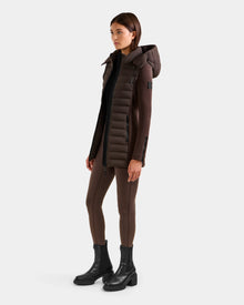 A woman faces slightly left, wearing Rudsak's MARRY M - L125519 Espresso long fitted brown puffer coat with down insulation and removable hood, brown leggings, and black ankle boots against a plain light background.