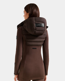 A woman with straight brown hair is seen from behind wearing the Rudsak MARRY M - L125519 Espresso long, fitted women’s hybrid jacket with a padded hood. The black Rudsak logo accents the hood and upper back against a plain light background.