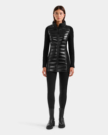 A woman faces forward wearing the Rudsak MARRY S - L125521 Black long, weather-resistant sleeveless puffer vest over a black long-sleeve top, leggings, and ankle boots against a plain light background.