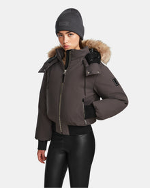 A person in a Rudsak MAUDE - L124522 Grey winter jacket with a fur-trimmed hood, gray beanie, and black faux leather pants stands against a plain white background.