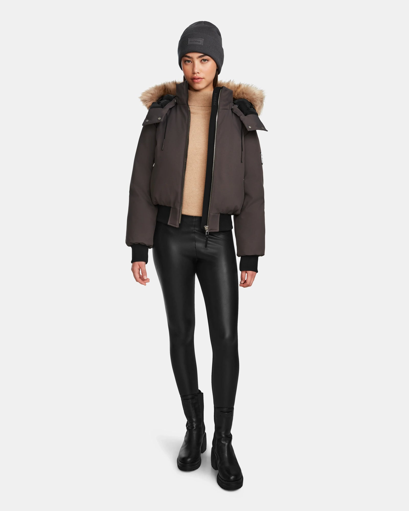 A woman stands against a white background wearing the MAUDE - L124522 Grey winter jacket by Rudsak, styled with a gray beanie, tan turtleneck, black faux leather leggings, and boots.