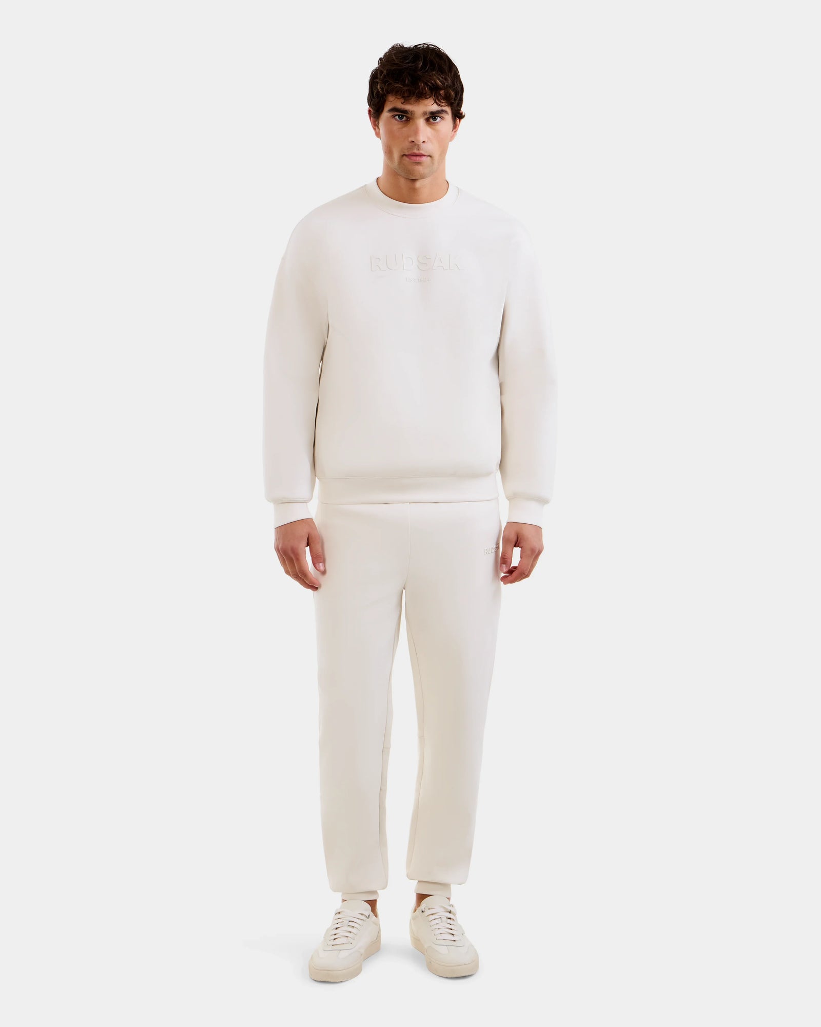 A man faces forward in the Rudsak MAXTON - U525007 Vanilla M jogger set—long-sleeve sweatshirt and matching pants with white sneakers—against a plain white background, perfect for everyday comfort.