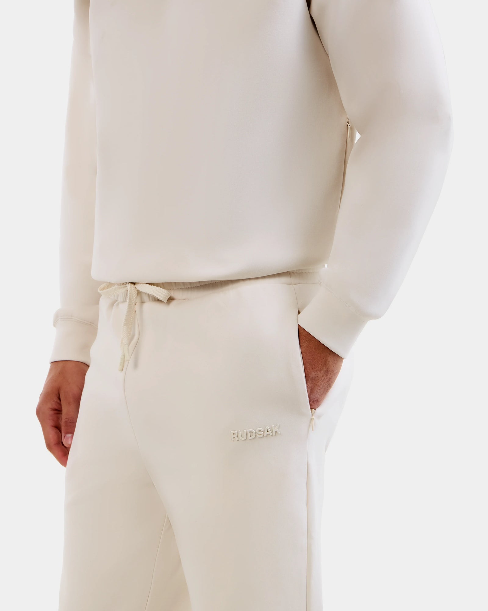 Someone wears the Rudsak MAXTON - U525007 jogger set in Vanilla M with a hand in the pocket. The cream-colored cotton-blend pants have "RUDSAK" embroidered near the pocket, providing everyday comfort against a clean white background.