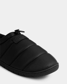 Close-up of Rudsak’s MILTON - M225504 Black men’s quilted slipper with textured rubber sole, faux-shearling lining, adjustable toggle on top, and a rectangular patch with raised lettering on a plain light gray background.