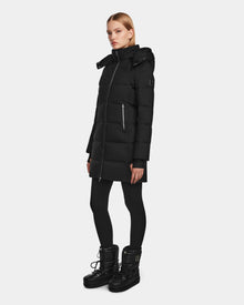 A woman wears the Rudsak MIRIAM - L123544 Black women’s down puffer coat, paired with black leggings and winter boots, standing slightly to the side against a plain light background.
