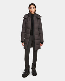 A woman stands facing forward, wearing the Rudsak MIRIAM - L123544 Grey women’s down puffer coat with a weather-resistant shell, paired with black pants and black winter boots against a plain white background.