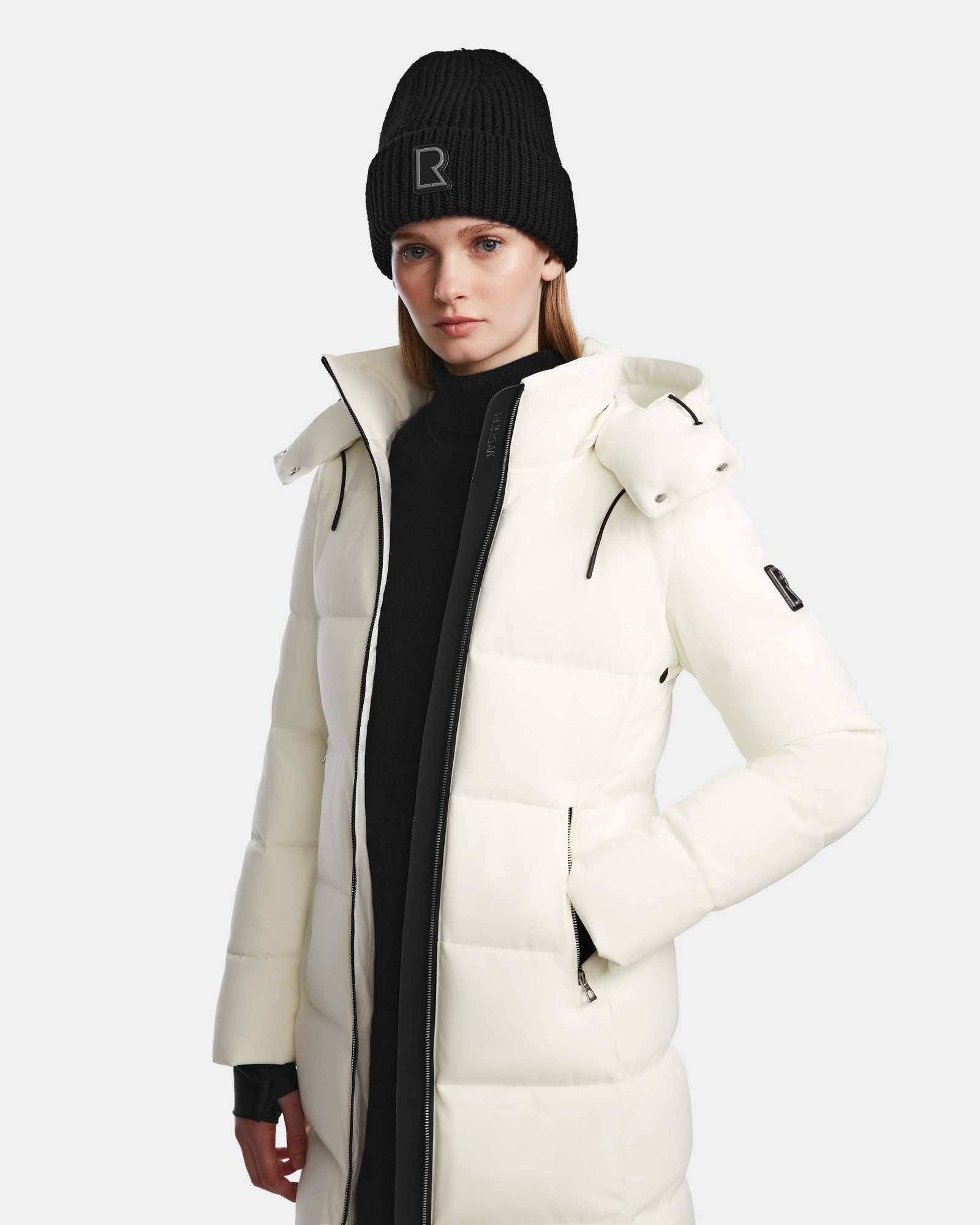 A woman wearing the Rudsak MIRIAM - L123544 Iceland white down puffer coat and a black beanie with an R stands against a plain light background, highlighting stylish winter essentials.