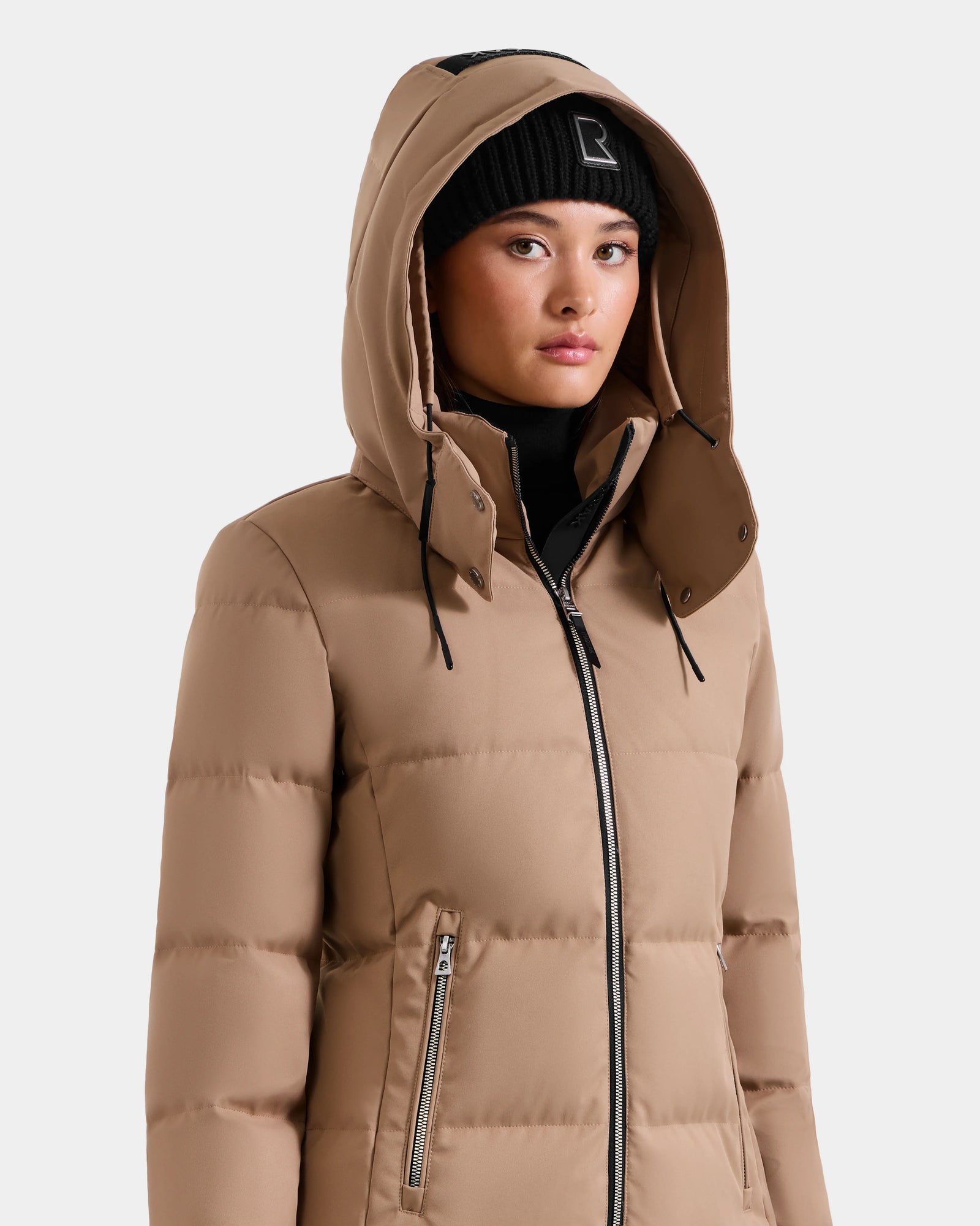 A person wearing the Rudsak MIRIAM - L123544 Sand women’s down puffer jacket with a hood and a black beanie looks slightly to the side. The weather-resistant jacket has zippered pockets and a high collar, set against a plain light gray background.
