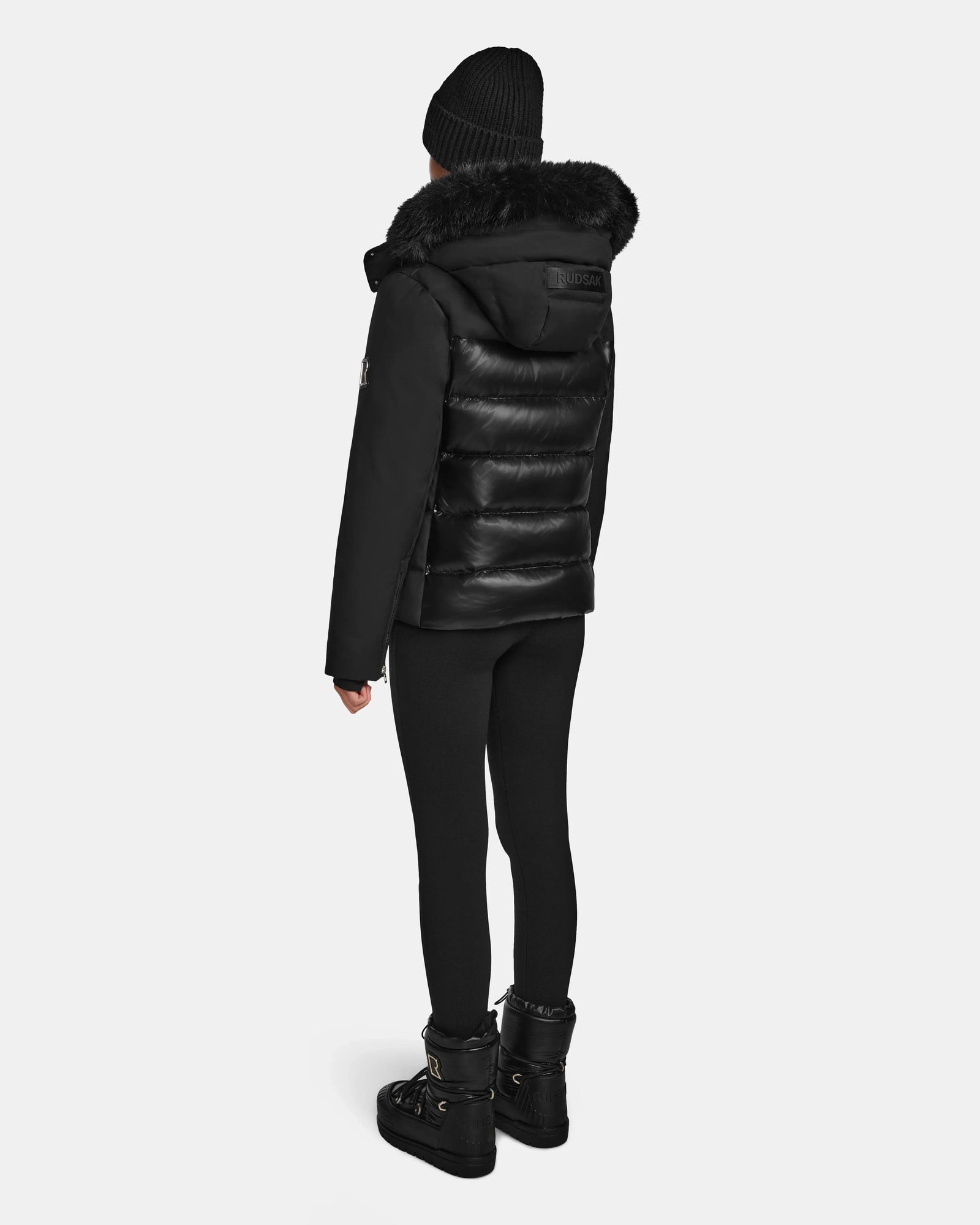 A person stands facing away, wearing the Rudsak MIRRY M - L124533 Black RDS-certified down winter coat with a fur-trimmed hood, paired with black leggings and boots, set against a plain light background.