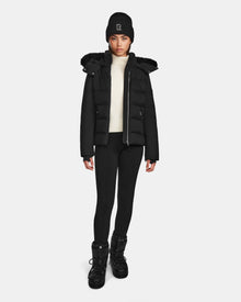 A person stands facing forward wearing the Rudsak MIRRY M - L124533 Black women's quilted down coat with a fur-trimmed hood, paired with a black beanie, white turtleneck, black leggings, and winter boots against a light background.