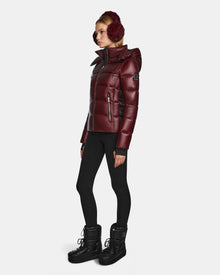 A woman in a shiny wine Rudsak MIRRY S - L123545 womens quilted down winter coat, matching earmuffs, black leggings, and black boots stands against a plain white background.