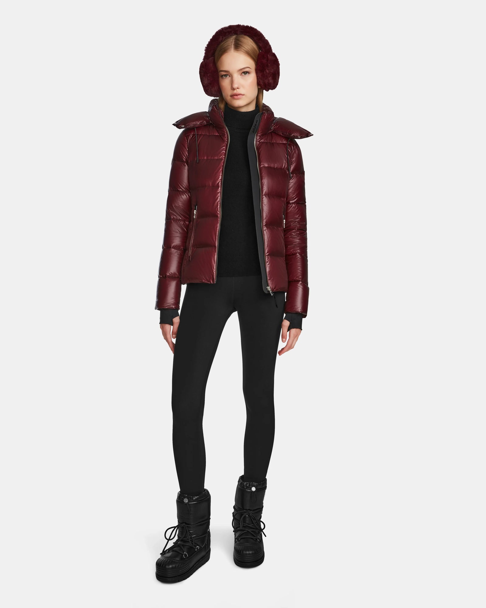 A woman stands against a plain white background wearing the Rudsak MIRRY S - L123545 Wine women’s quilted down coat, matching earmuffs, black leggings, a black turtleneck, and black winter boots.