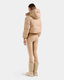A person stands facing away, wearing Rudsak's OPRAH - L125546 Vanilla RDS-certified women’s cropped puffer jacket with a hood, paired with form-fitting beige leggings and beige knee-high fur-trimmed boots, against a plain white background.