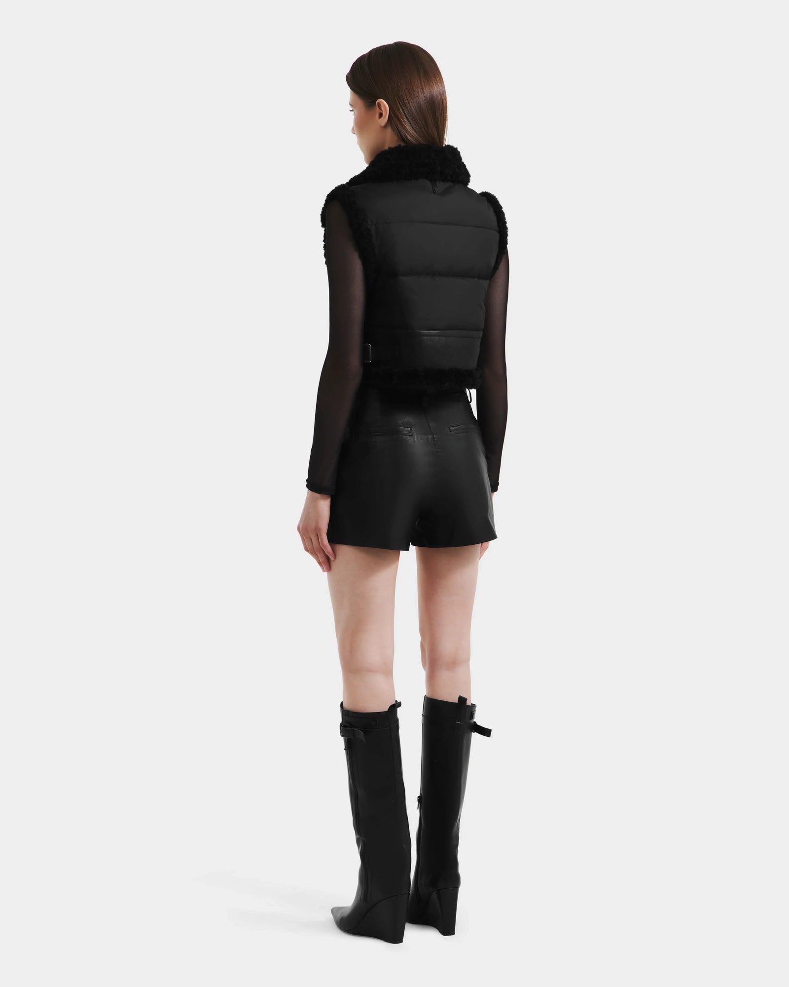 A woman faces away, wearing Rudsak's PAISLEY - L125516 Black vegan leather vest over a sheer top, paired with black leather shorts and knee-high black boots, set against a plain white background.