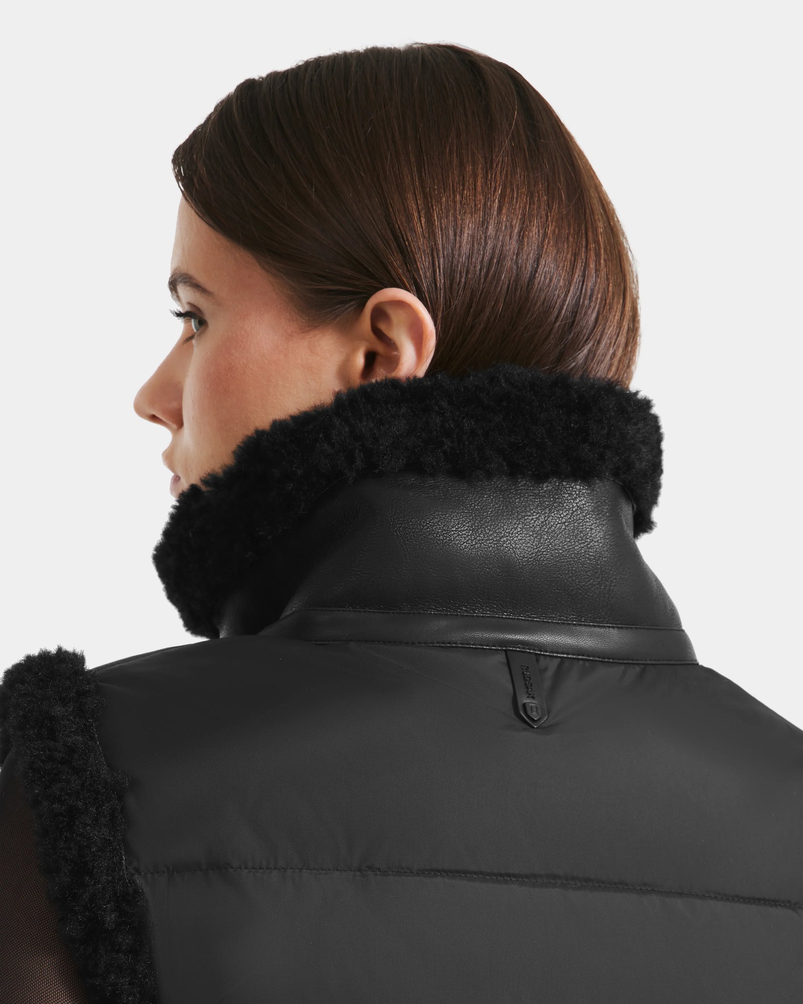 A person with straight brown hair is seen from behind, wearing the Rudsak PAISLEY - L125516 Black jacket featuring a textured faux shearling collar, set against a plain, light-colored background.