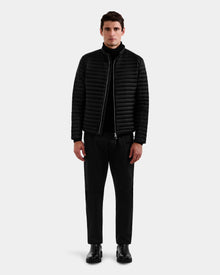 A man stands facing forward wearing the Rudsak REYMOND M - M125523 black men’s down jacket made from recycled nylon with RDS-certified down, paired with a black turtleneck, pants, and boots against a plain light gray background.