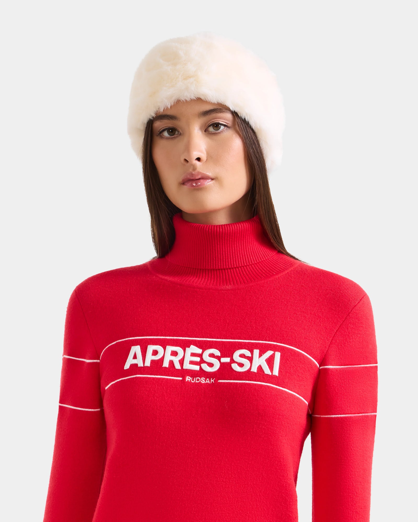A woman wears the SANDIE - L425552 Iceland faux fur headband by Rudsak and a bright red turtleneck sweater with APRÈS-SKI across the chest, standing against a light gray background—a chic winter accessory look.