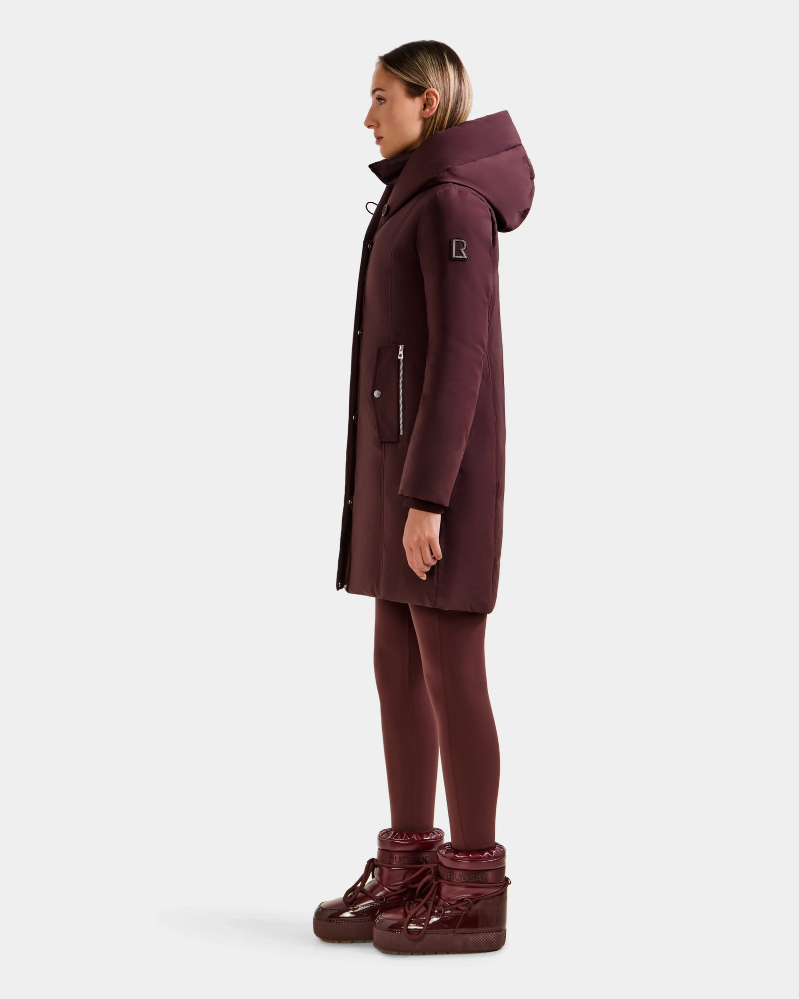 A woman stands in profile wearing the Rudsak SARINA - L125550 Bordeaux down parka with matching maroon leggings and maroon winter boots against a plain light background.