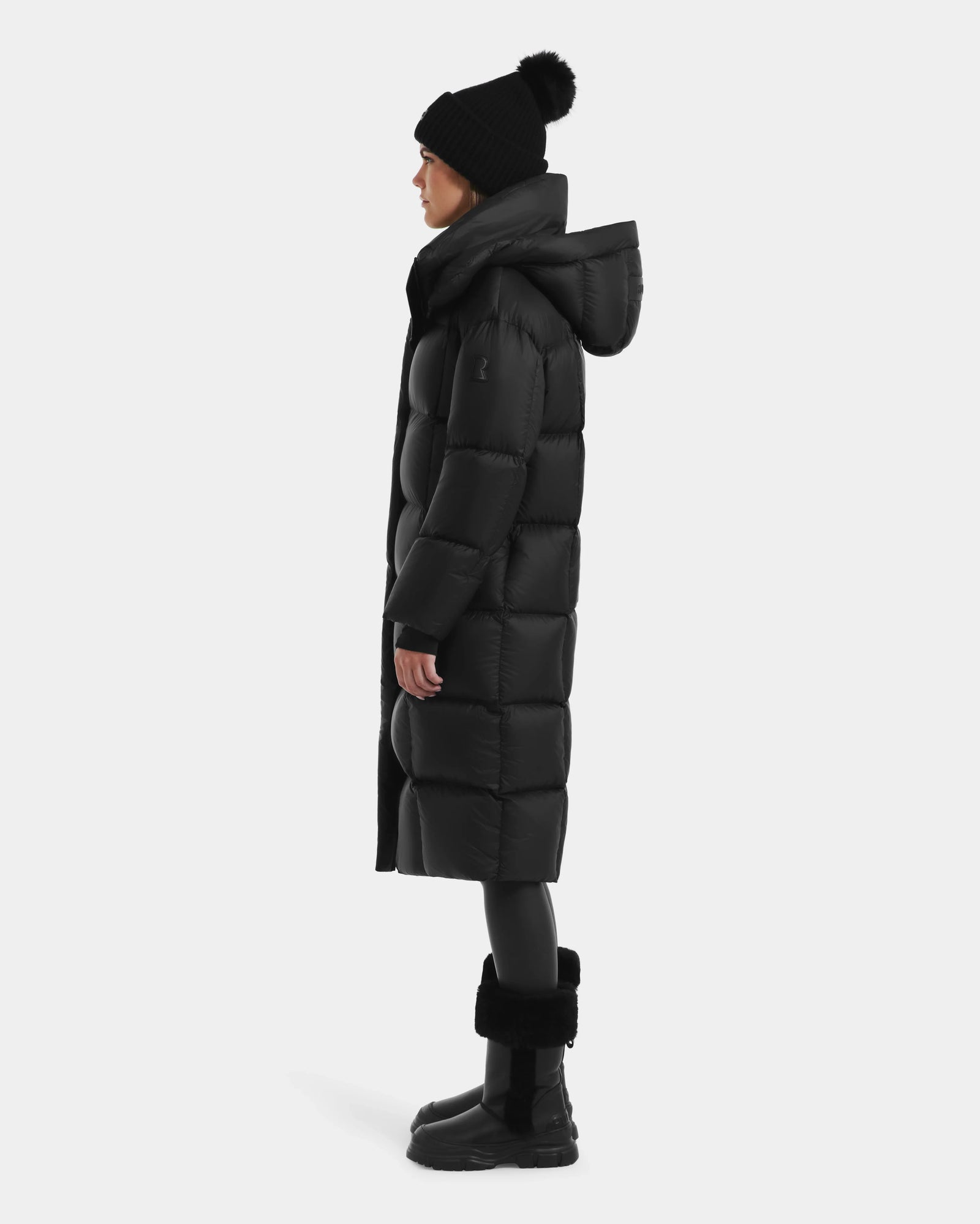 A person in the Rudsak SASHA - 8125686 Black women’s long down puffer coat with removable hood, black beanie with pom-pom, leggings, and boots stands in profile against a plain light background.