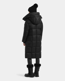 A person wears the Rudsak SASHA - 8125686 Black women’s long down puffer coat with removable hood, paired with black leggings, fur-trimmed boots, and a black beanie, standing against a plain light gray background.