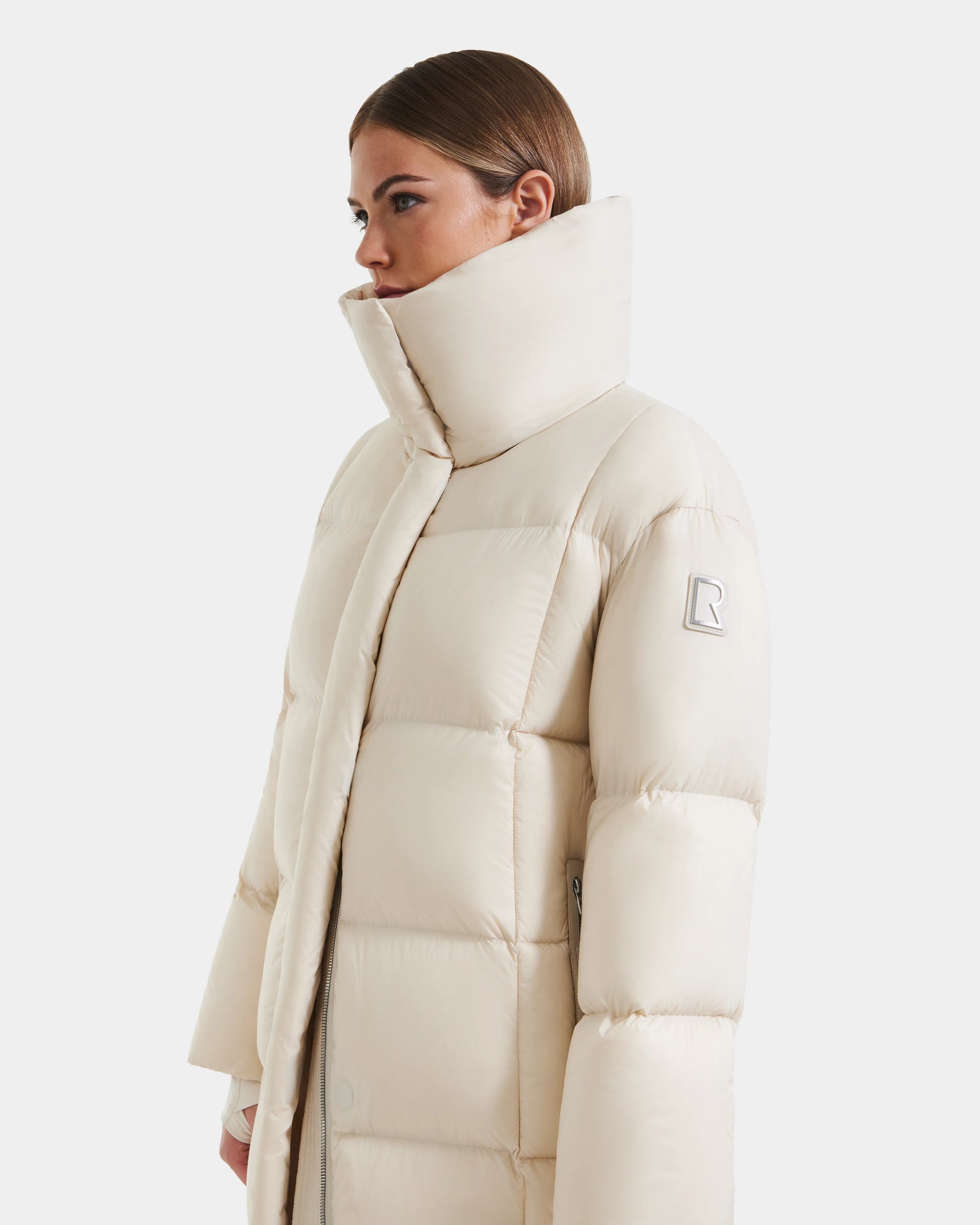 A woman with brown hair in a low bun wears the Rudsak SASHA - 8125686 Vanilla long down puffer coat, featuring a high collar and logo patch on the sleeve, standing against a plain white background.