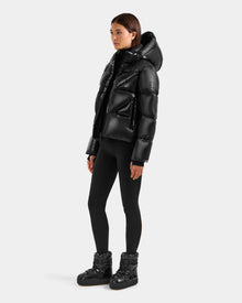 A person stands against a plain light gray background wearing the Rudsak STEFANIE S - L125585 Black quilted down puffer with a hood, paired with black leggings and black padded ankle boots.