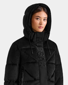 A woman wears the Rudsak STEFANIE V - L125584 black down puffer jacket, featuring the brand's name vertically on the front, as she stands against a plain light gray background.
