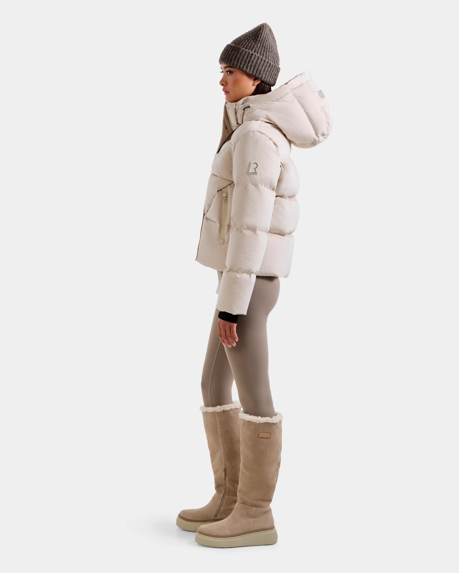 A person stands in profile wearing the Rudsak STEFANIE V - L125584 Vanilla down puffer jacket, paired with taupe leggings, tall beige shearling boots, and a knit beanie against a plain white background.