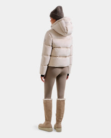 A person stands facing away in the Rudsak STEFANIE V - L125584 Vanilla beige down puffer jacket with hood, paired with taupe leggings, tan knee-high shearling boots, and a brown knit beanie against a light grey background.