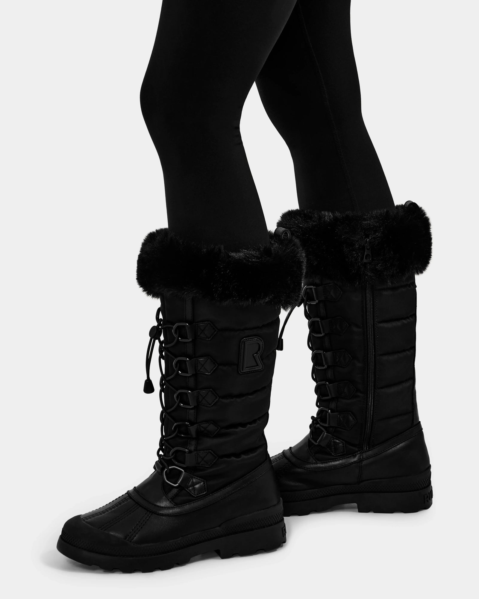 A person wearing Rudsak WINDY - L224507 Black women's winter boots with faux fur trim and lace-up fronts, paired with black leggings, stands against a plain light background.