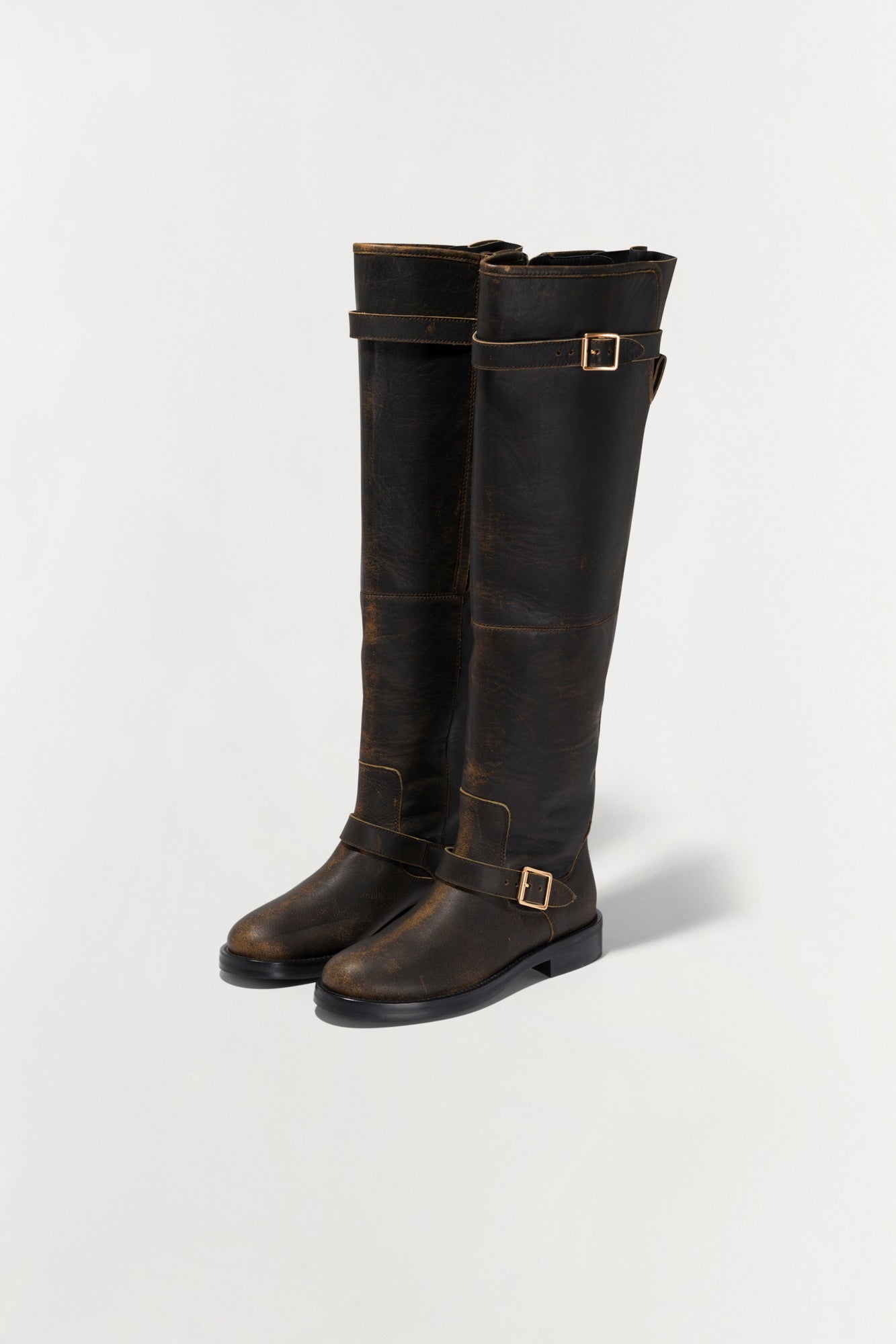 Black and brown distressed leather over-the-knee boots with gold buckle straps and black soles.