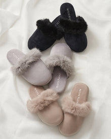 A collection of Petite Plumes Womens Faux Fur Trim Slippers, featuring three plush slip-on pairs in black, gray, and beige with elegant fur trim, displayed on a white fabric background.
