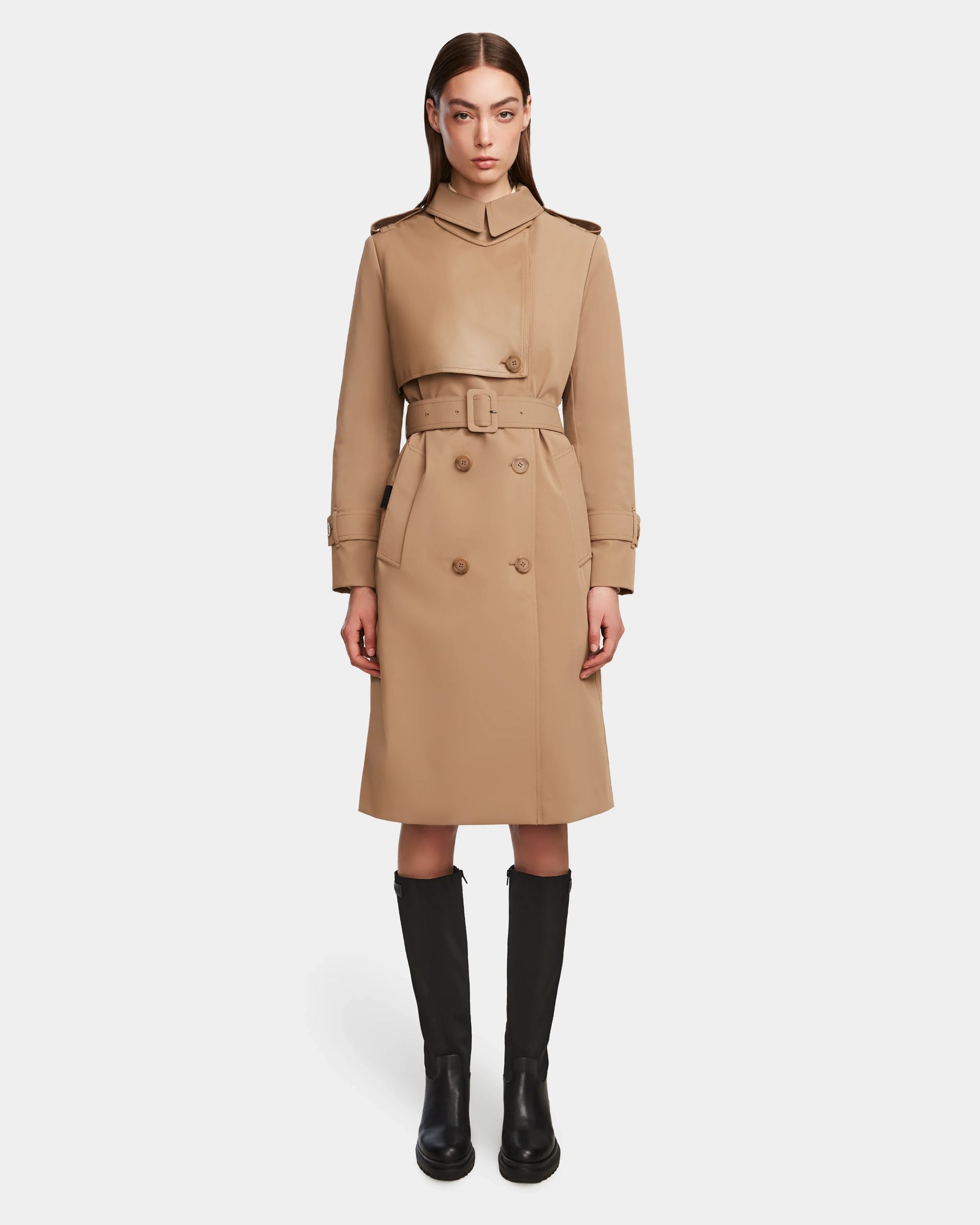 A person faces forward against a plain white background, wearing the Rudsak KINDA - 8124690 Camel womens trench coat with a belt and black knee-high boots.