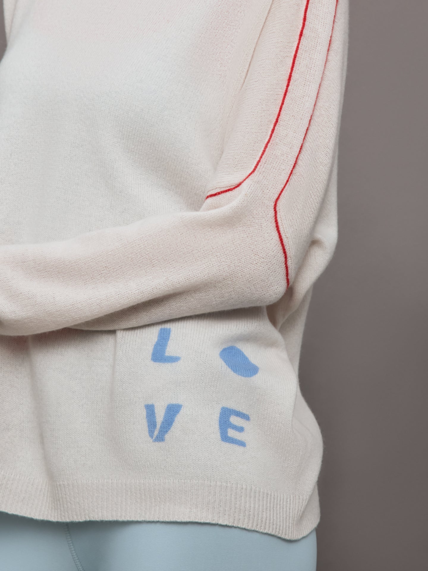 Model wears a white sweater with red seams along the biceps and blue block lettering on front bottom right that says LOVE. Back of sweater says in blue lettering "Love is all around you."