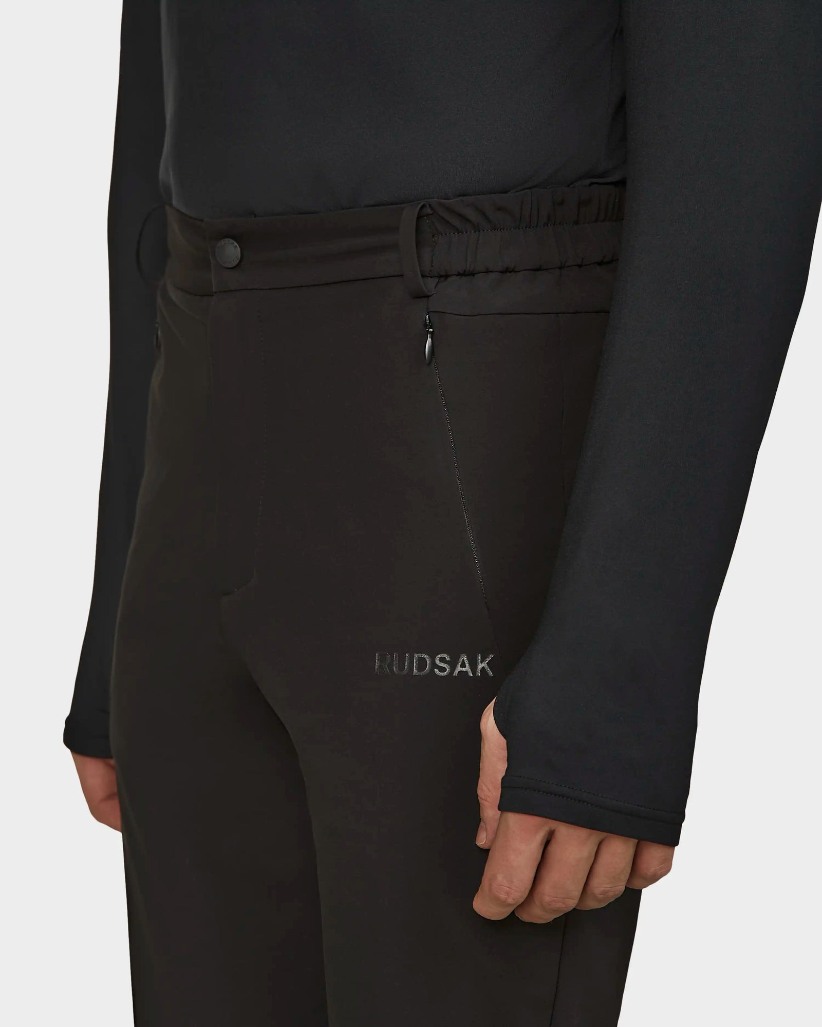 A person wears Rudsak KICO - 6523508 Black tailored pants with a zippered pocket and a black long-sleeve top with thumb holes. Only the torso and upper legs are shown, emphasizing the sleek men's outdoor pants style.