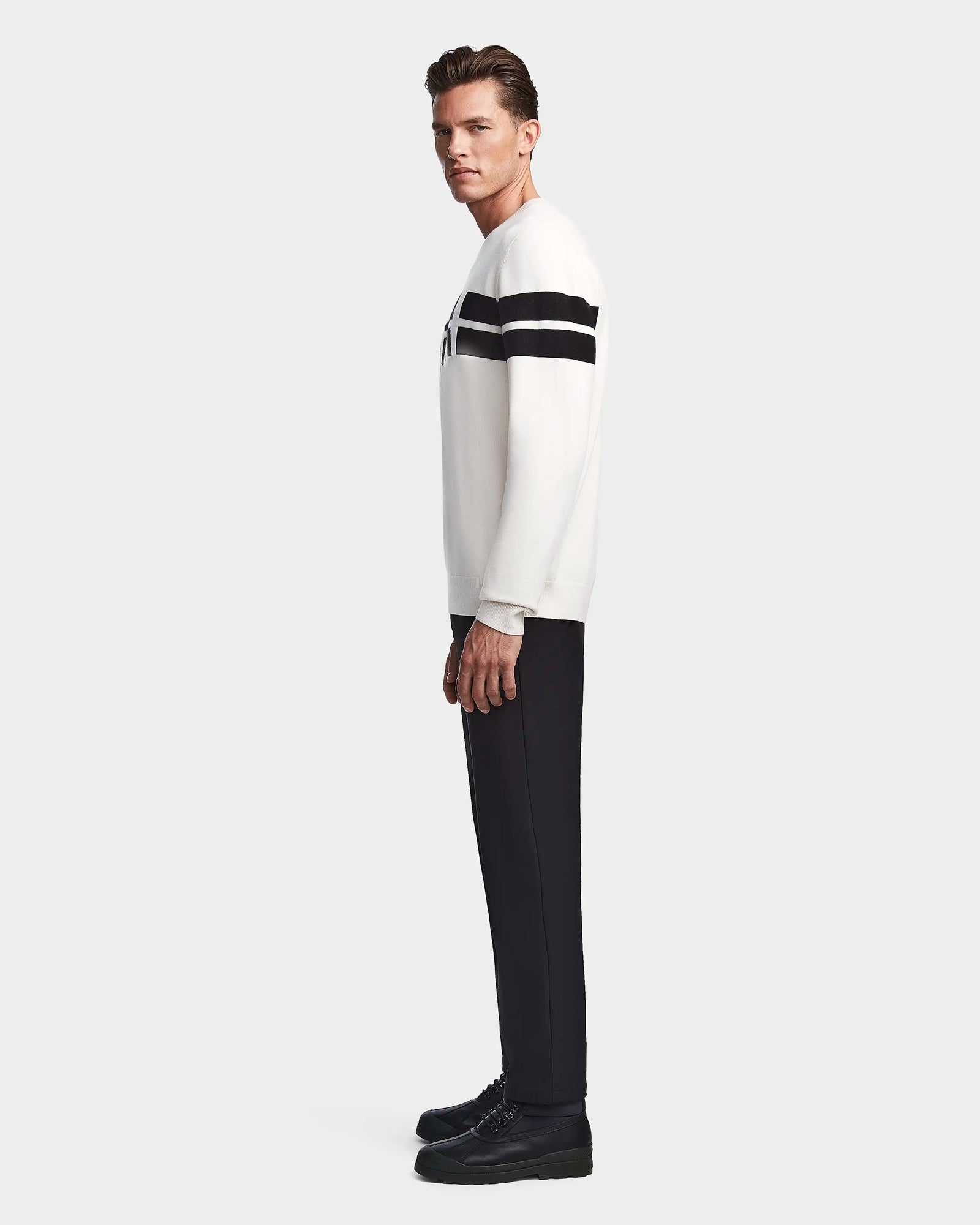 A man stands in profile against a plain background, wearing the Rudsak OMAR - 6523507 Iceland men’s knit sweater with black horizontal stripes on the upper sleeves, paired with black pants and shoes.
