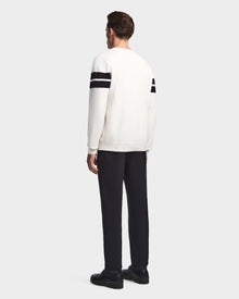 A man faces away, wearing the Rudsak OMAR - 6523507 Iceland white men’s knit sweater with two black stripes on each sleeve, paired with black pants and shoes, against a plain light gray background.