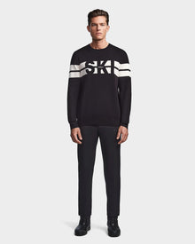 A man faces forward wearing the Rudsak OMAR - 6523507 Black mens knit crewneck sweater with white sleeve stripes and SKI on the chest, paired with black pants and shoes, set against a plain light background.