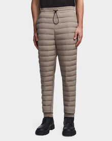 A person wears Rudsaks MIKE - 6523503 Cement mens quilted pants with a drawstring waist, paired with a black shirt and lace-up boots. These insulated pants provide warmth and style against a white background.
