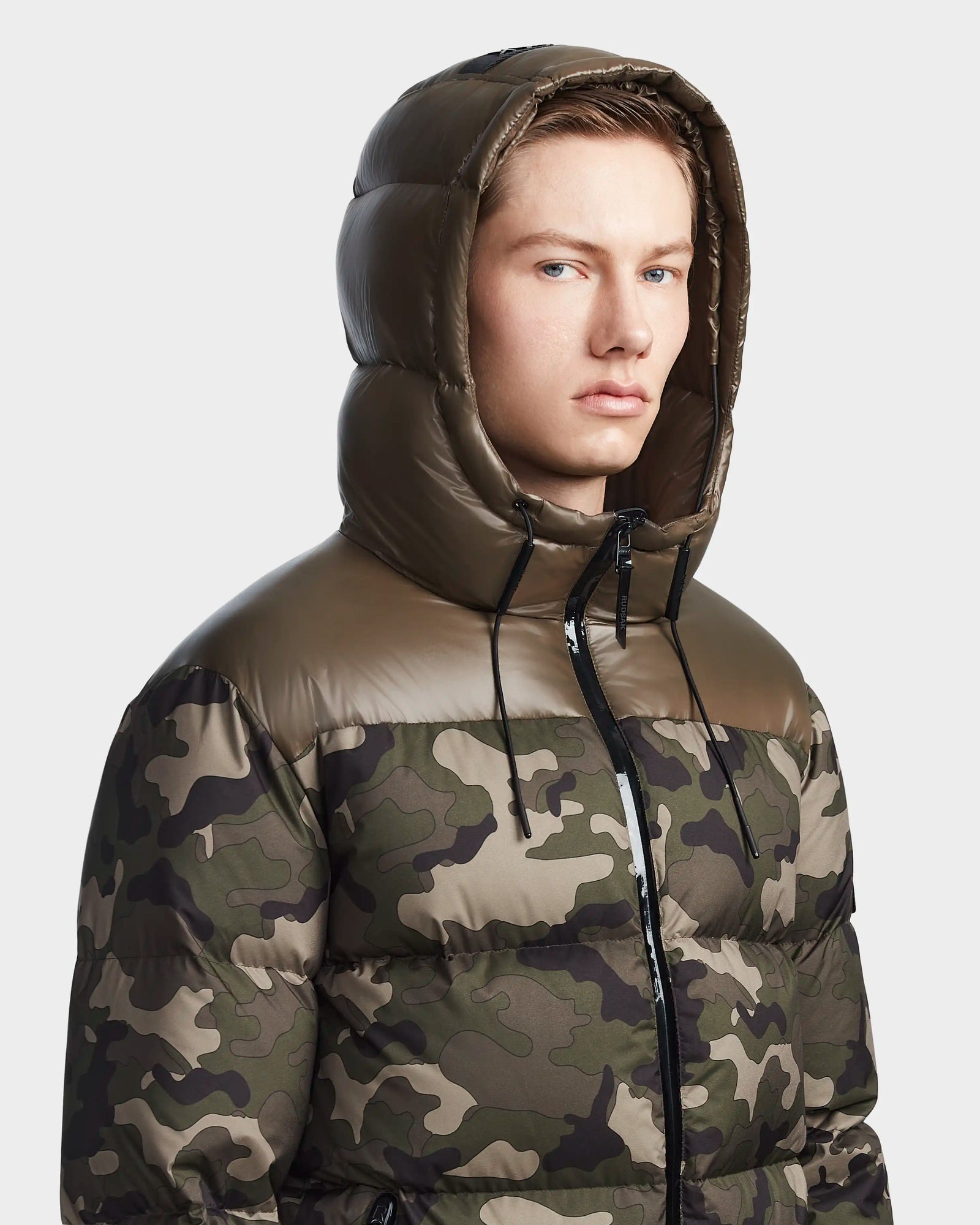 A young man models the RONNY M - M123512 Camouflage men’s hooded down puffer by Rudsak, featuring green and brown camouflage and a shiny hood, as he looks slightly to the side against a plain light background.