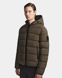 A young man with straight brown hair wears the Rudsak DEMIAN M - M123507 Light Olive puffer jacket, made from sustainable recycled fabric with a hood and black zippers, standing against a plain white background.