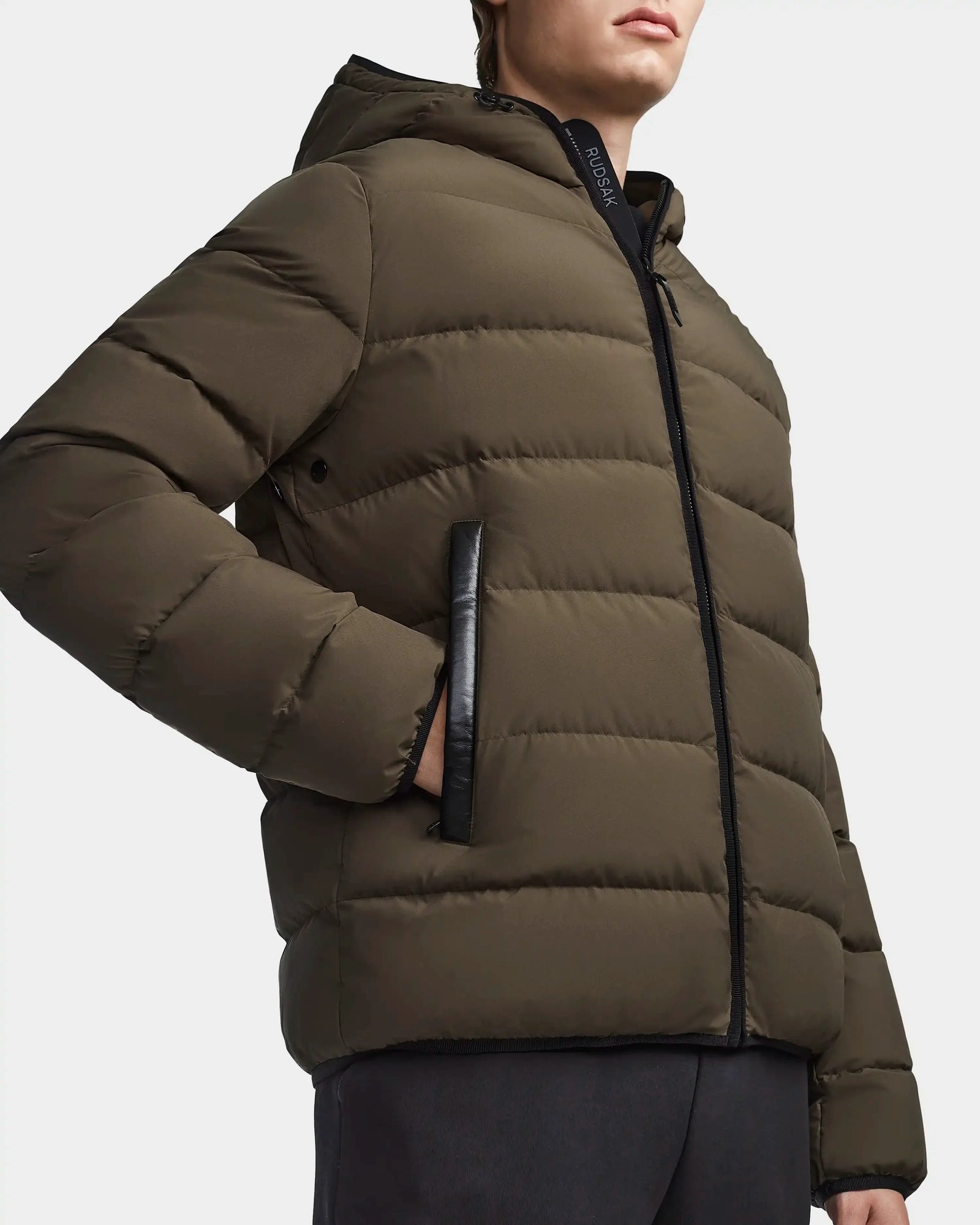 A person wears the Rudsak DEMIAN M - M123507 men’s down puffer in light olive with black trim and hood, crafted from recycled fabric. Standing against a plain background, they have a hand tucked into one zippered pocket.