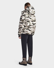 A person stands facing away, wearing the Rudsak VINCENT M - 6123510 Camouflage men's down puffer in white, gray, and black camo with a hood, black pants, and tan boots against a plain light gray background.