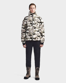 A person stands facing forward, wearing the Rudsak VINCENT M - 6123510 Camouflage men's down puffer in beige and gray camo, paired with black pants and tan lace-up boots, against a plain light gray background.