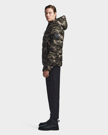 A person stands in profile wearing the Rudsak DEMIAN M-R - M123504 Camouflage mens down puffer hooded jacket, paired with black pants and black boots, against a plain white background.