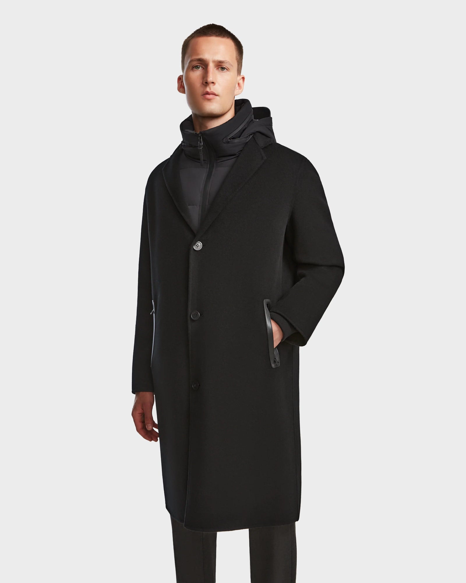 A man wearing the Rudsak DRAL - 6124503 Black mens long wool jacket with a high-neck inner layer stands against a plain white background, gazing slightly upward.